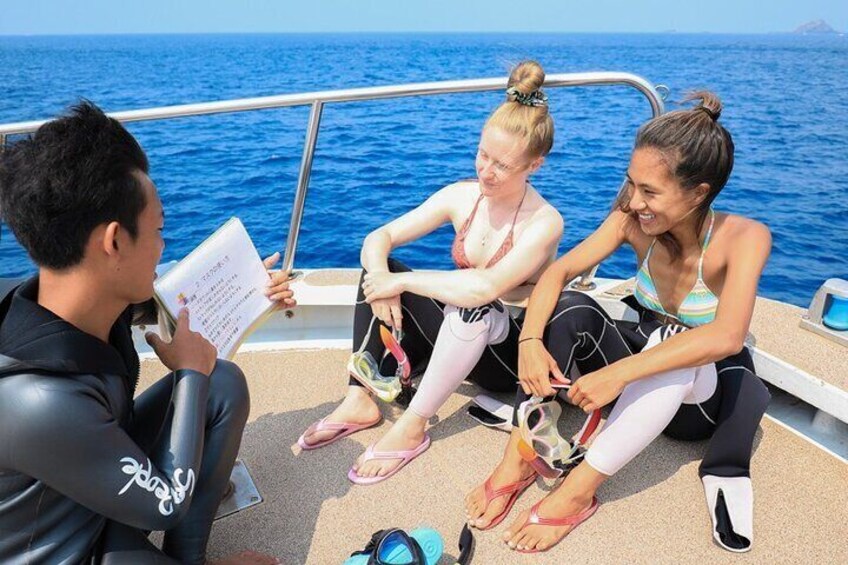 Three people on a boat, one reading a document, with a clear blue sea and a distant island in the background. 5