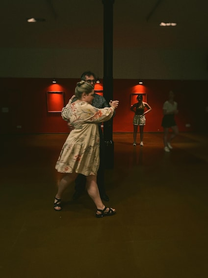 Une personne vêtue d’une robe à motifs danse avec un partenaire dans une pièce faiblement éclairée, aux murs rouges et ornée d’une colonne. 4