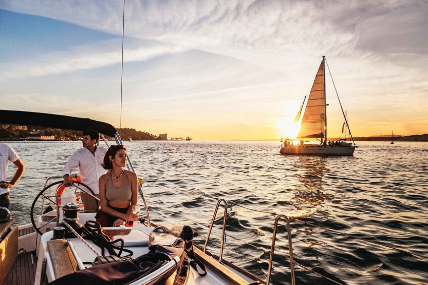 A sailboat with people on board, a sailboat in the background, and a sunset over the water. 1
