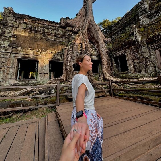 A person standing on a wooden platform in front of ancient stone ruins with large tree roots intertwined with the structure. 4