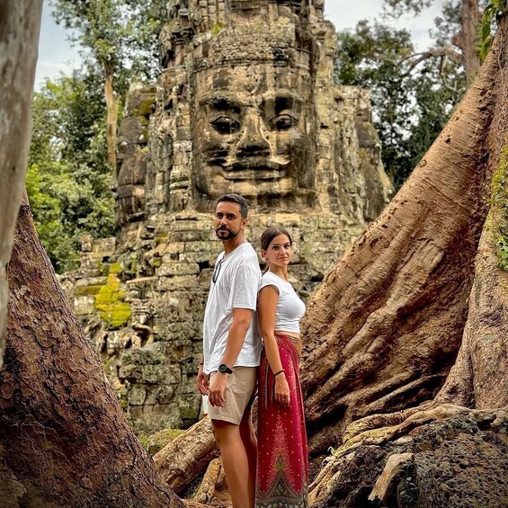 A man and woman standing in front of a large stone face structure surrounded by tree roots. 5