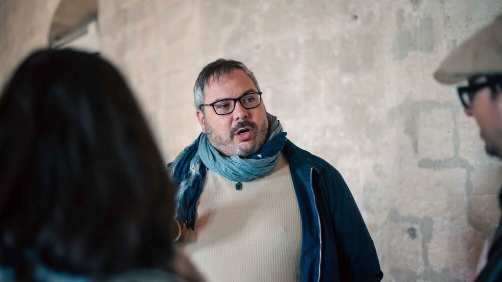 A man in a sweater and scarf, conversing with two others in a stone-walled room. 4
