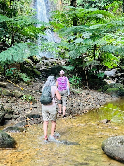 Two hikers crossing a shallow stream in a forested area with a waterfall in the background. 4
