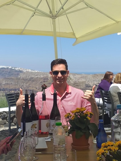 A man giving a thumbs-up at a table with wine bottles, glasses, and a potted plant. 2