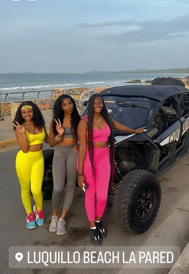 Three people posing by a vehicle on a beach with a rocky shoreline and ocean in the background. 5