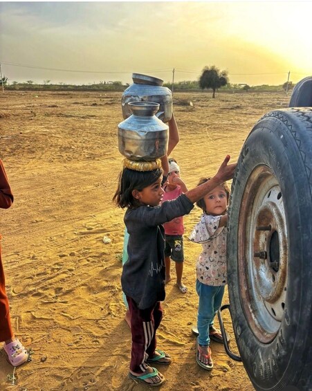 Children carrying metal pots on their heads, a large tire, and a person in the background. 4