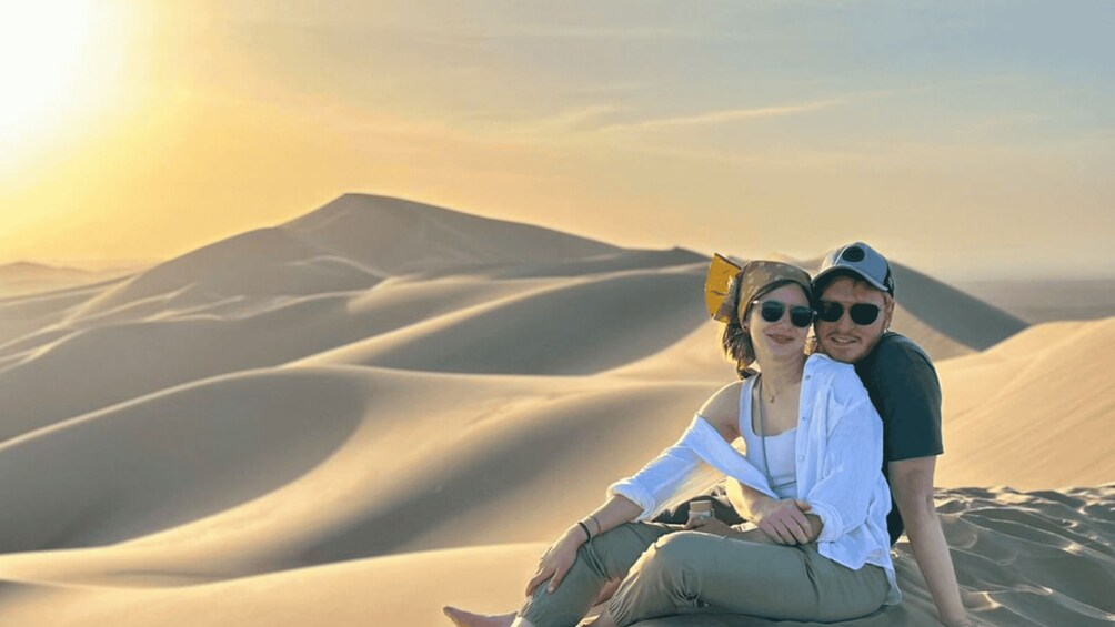 Two people sitting on sand dunes with a desert landscape in the background. 2