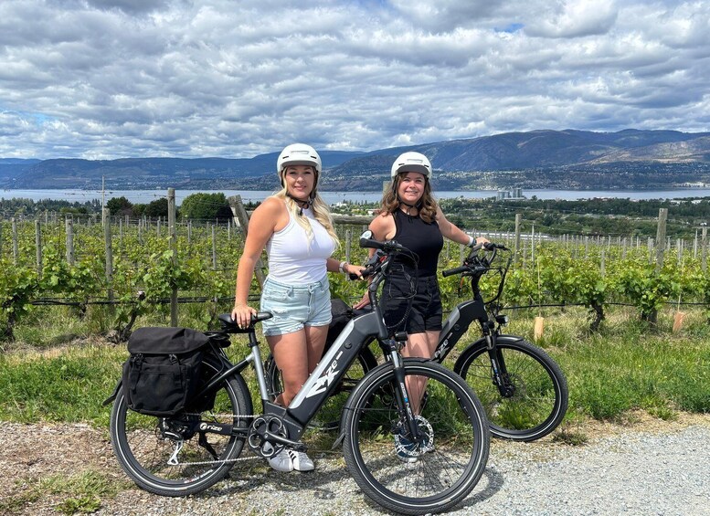Two people standing next to bicycles in a vineyard with a lake and mountains in the background. 1