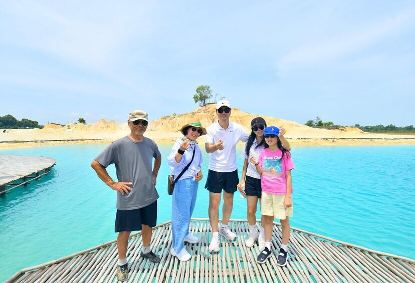 A group of people standing on a wooden dock by a turquoise body of water with a sandy beach and dunes in the background. 2
