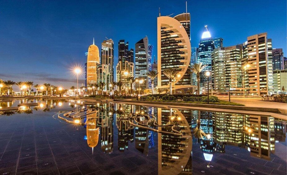 A city skyline at dusk with illuminated buildings and a reflective water feature. 2