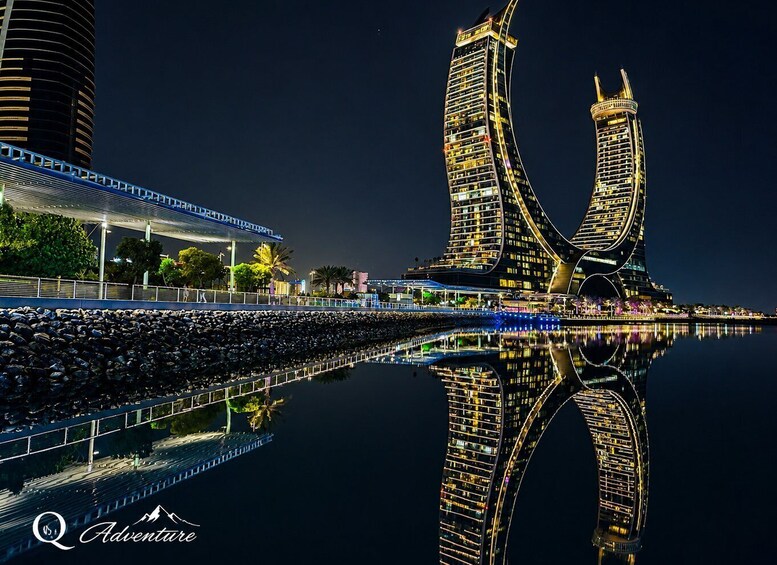 A nighttime cityscape with two illuminated skyscrapers reflecting on the water, a covered pedestrian walkway, and a bridge. 1