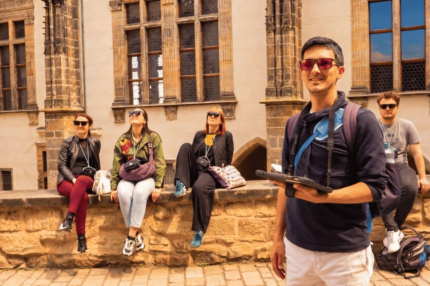 A group of people sitting on a stone wall in front of a historic building with large windows and intricate stonework. 2