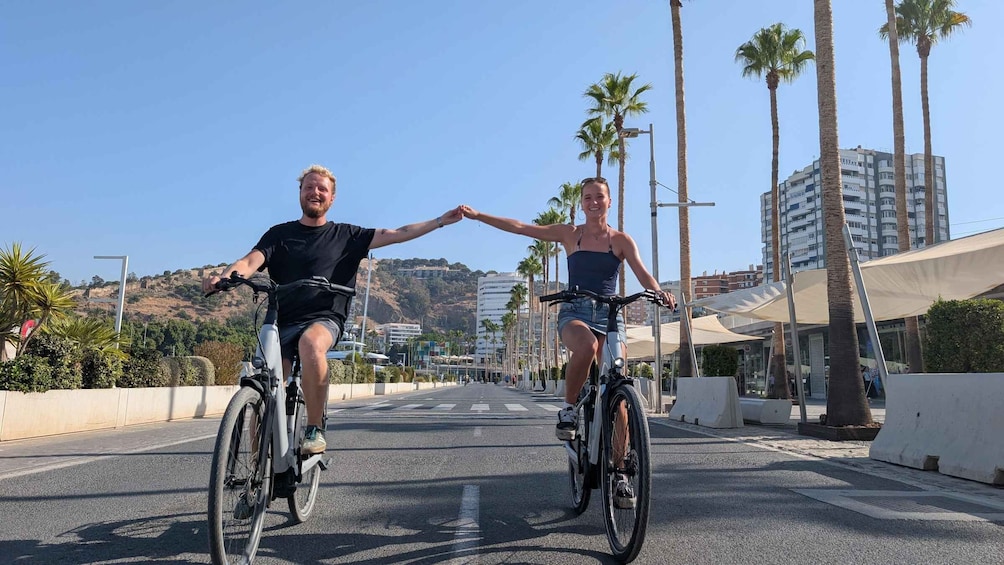 To personer cykler på en vej kantet med palmer og bygninger. 4