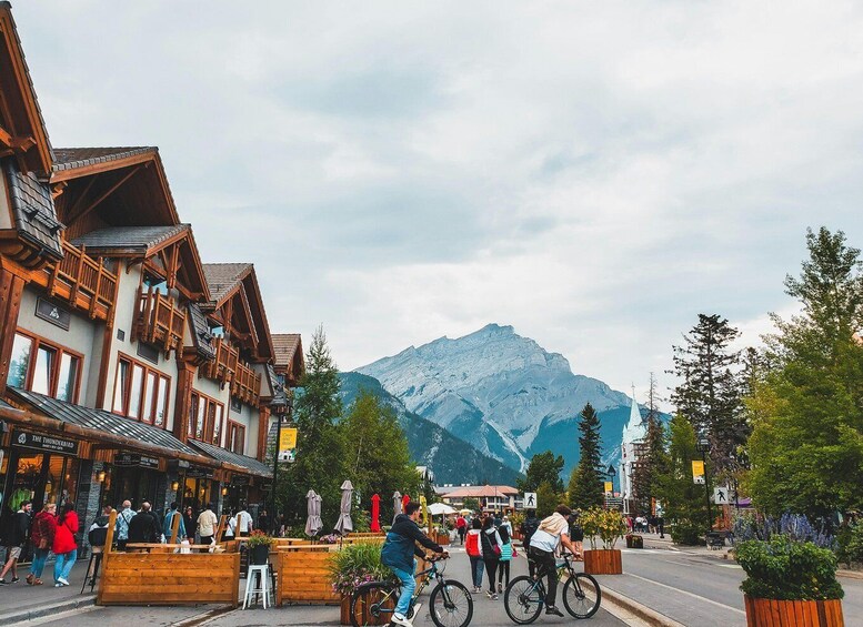 A mountain town with wooden buildings, cyclists, and a snow-capped mountain in the background. 1