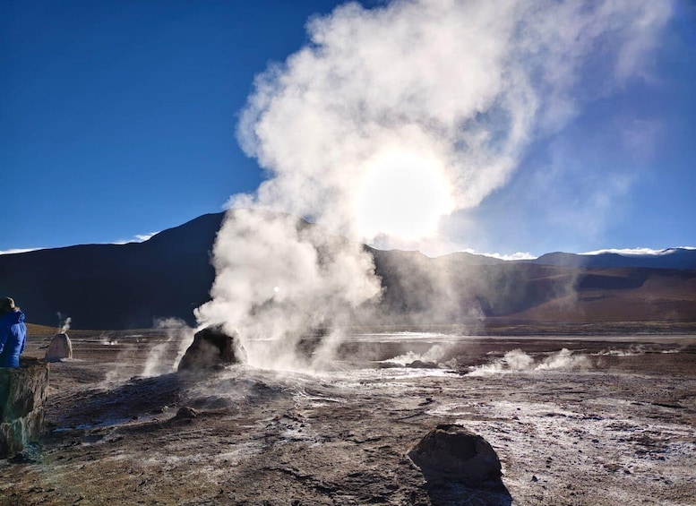 A geyser erupting with steam, a person observing, and a mountainous landscape in the background. 3
