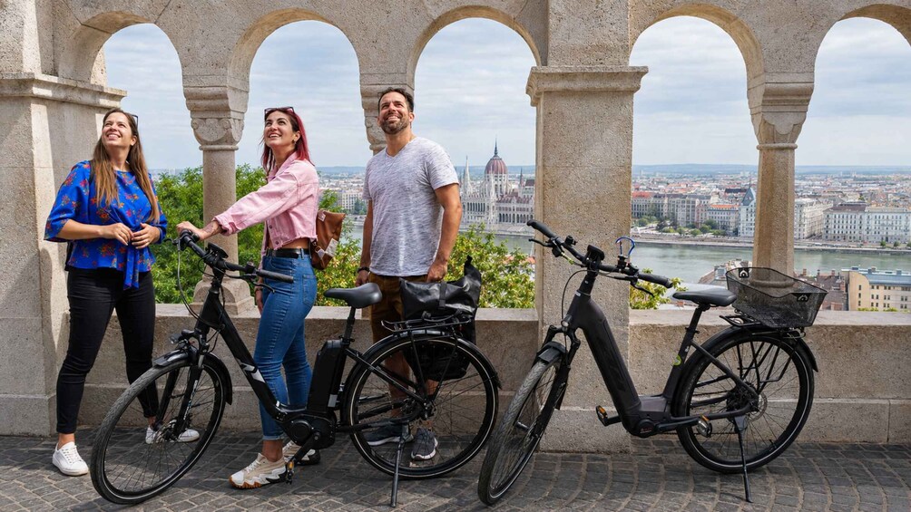 Three people standing next to bicycles, with a cityscape and river in the background. 1