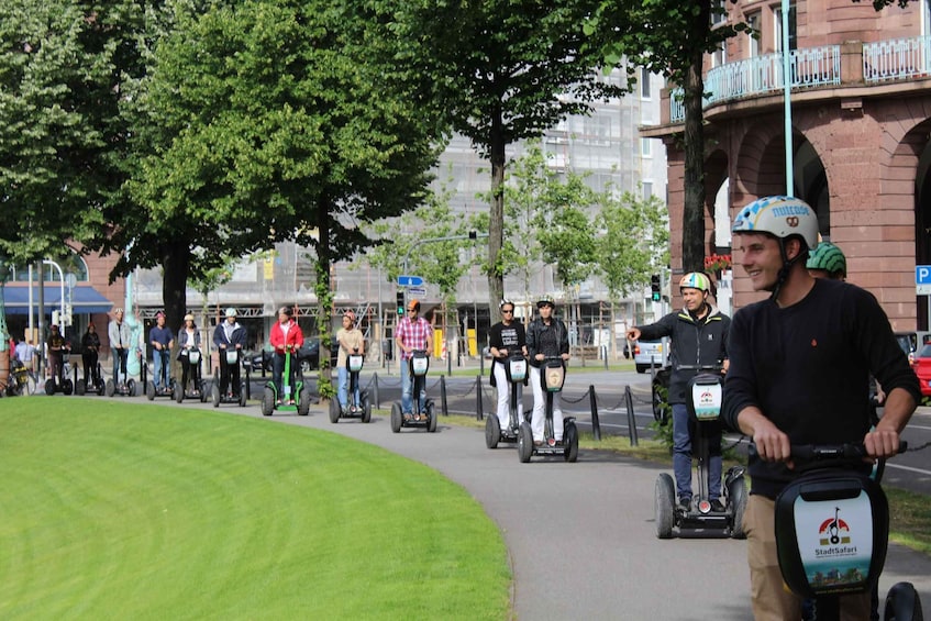 A group of people riding Segways on a paved path in an urban setting with trees and buildings in the background. 2