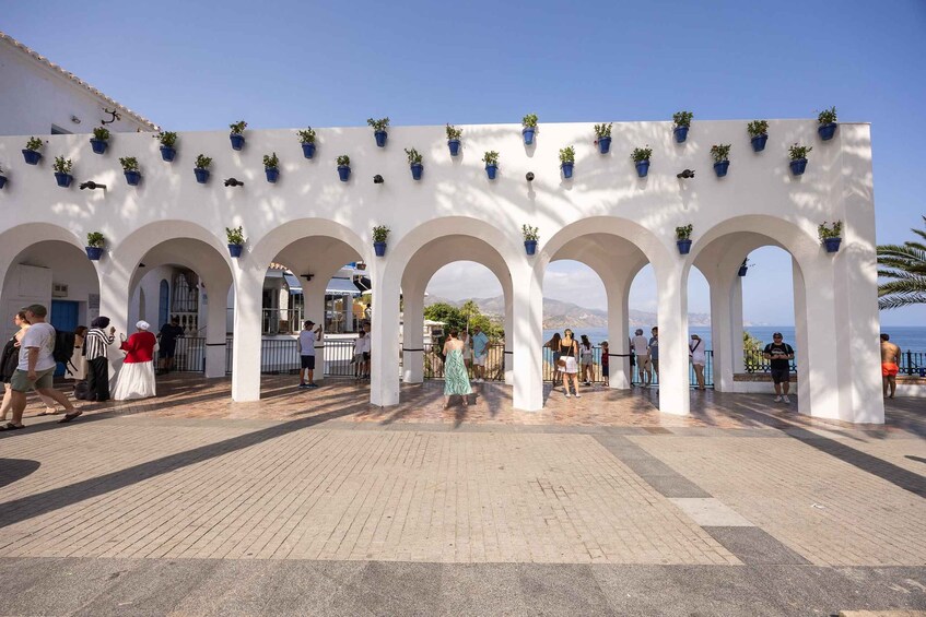 A covered walkway with arches and potted plants, overlooking a coastal area. 1