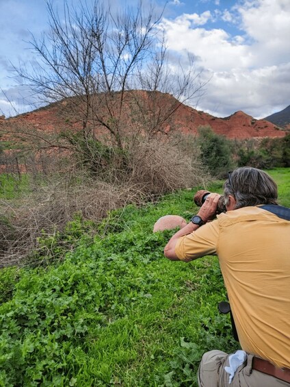 A person taking a photograph of a landscape with green vegetation and red hills in the background. 2