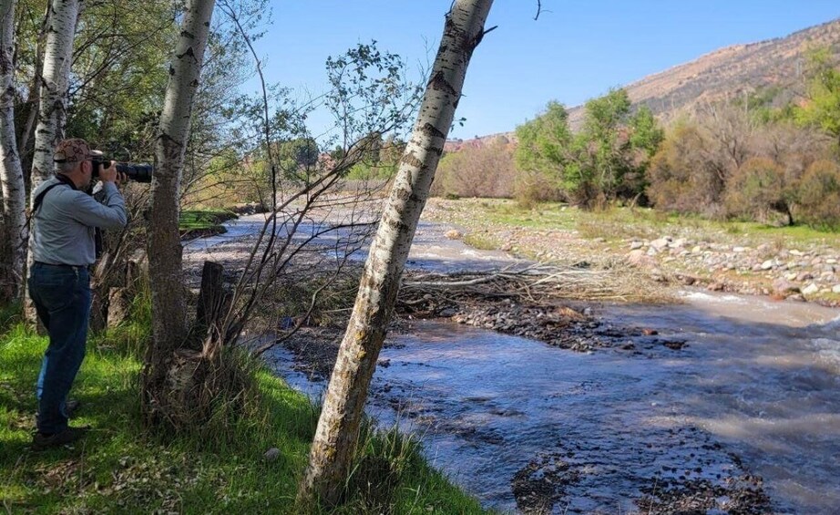 A person taking a photograph of a river with trees and a mountain in the background. 5