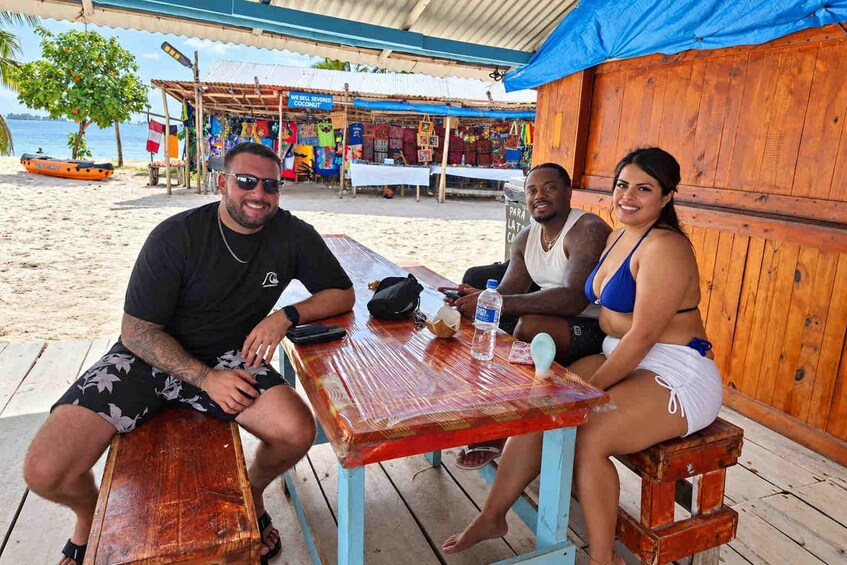 Three people sitting at a wooden table on a beach with a market in the background. 5