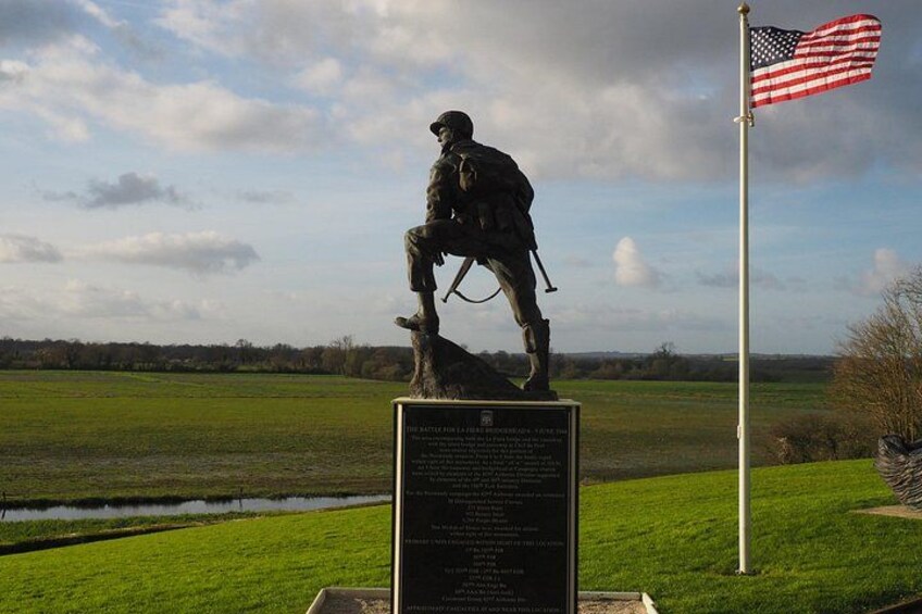 Una estatua de un soldado sobre un pedestal con una bandera estadounidense, ubicada en un paisaje rural con una masa de agua y árboles. 5
