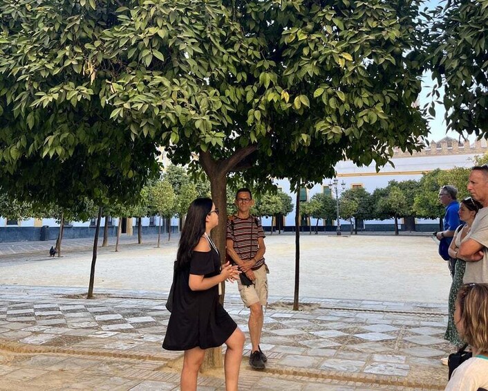 A group of people standing under a tree in a plaza with stone flooring and a blue sky in the background. 5