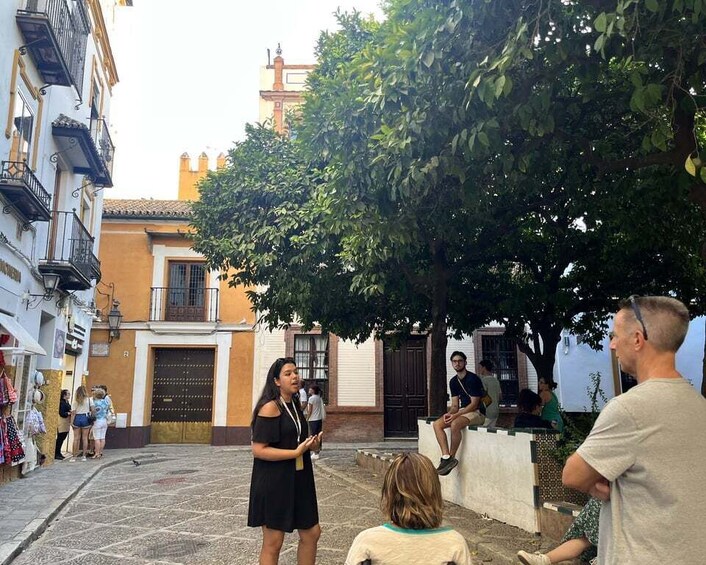 A cobblestone street with people walking and sitting, surrounded by buildings with balconies and a tree providing shade. 5