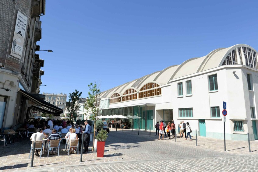 A café with outdoor seating, a modern building with arched windows, and people walking on the street. 2