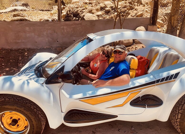 A couple in a small, white, open-top vehicle with yellow accents, parked outdoors. 4