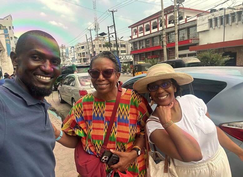 Three people standing on a street with buildings and cars in the background. 1