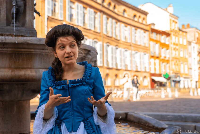 A person in historical costume standing in front of a fountain with classical architecture in the background. 1