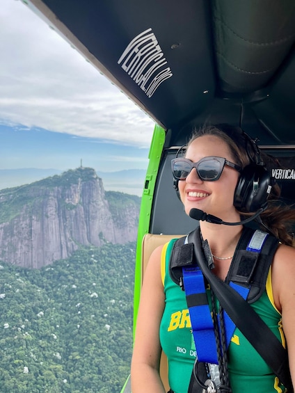 A person in a green and yellow outfit with a headset, seated in a helicopter cockpit, overlooking a mountainous landscape. 3