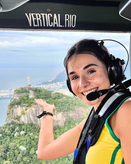 A person in a yellow top with headphones, standing in a helicopter cockpit with a view of a city and a mountain. 1