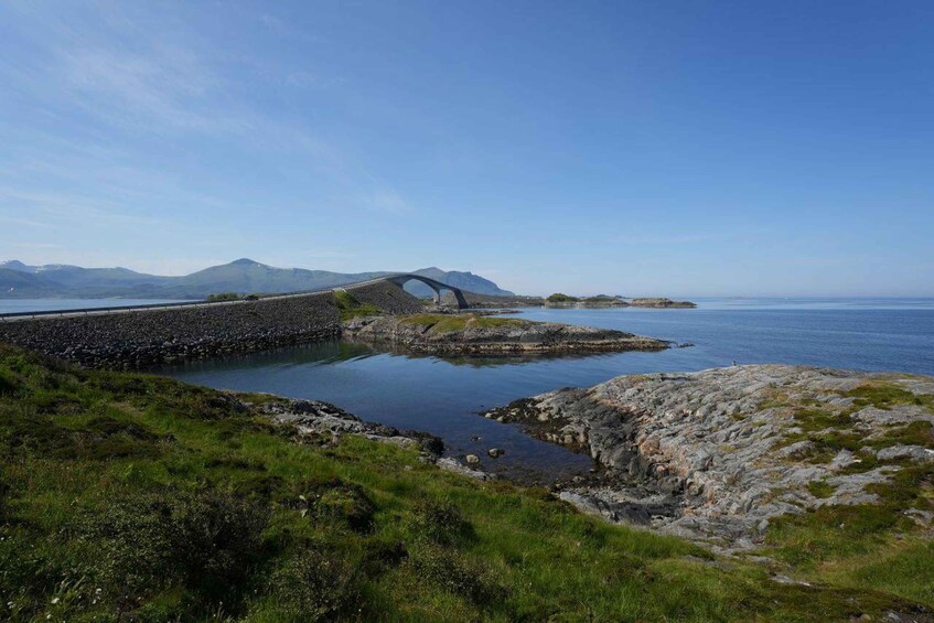 A coastal landscape with a stone bridge over a calm body of water, surrounded by rocky terrain and distant mountains. 1