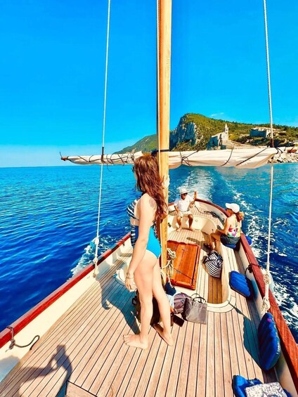 A sailboat with a woman in a swimsuit on deck, a wooden floor, and a clear blue sea with a distant rocky coastline. 3