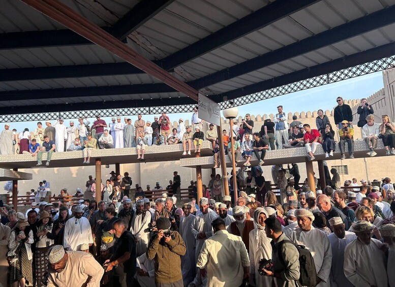 A crowded outdoor event with people standing and sitting on tiered platforms, under a covered structure. 2