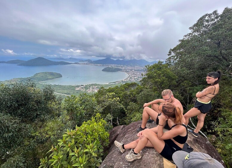 A group of people sitting on a rock overlooking a coastal landscape with mountains and a town in the distance. 4