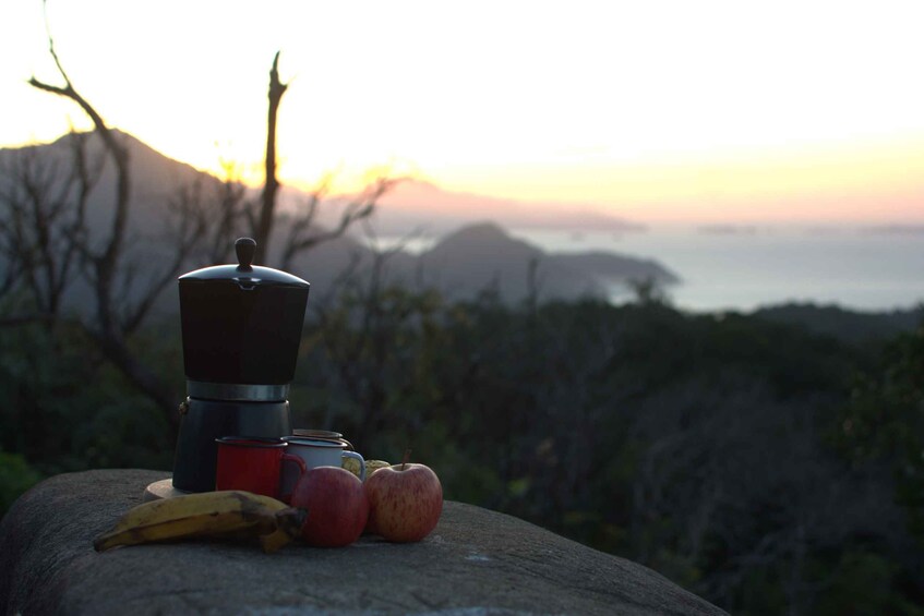 A portable coffee maker, a red mug, bananas, and apples on a rock with a sunset and mountains in the background. 5