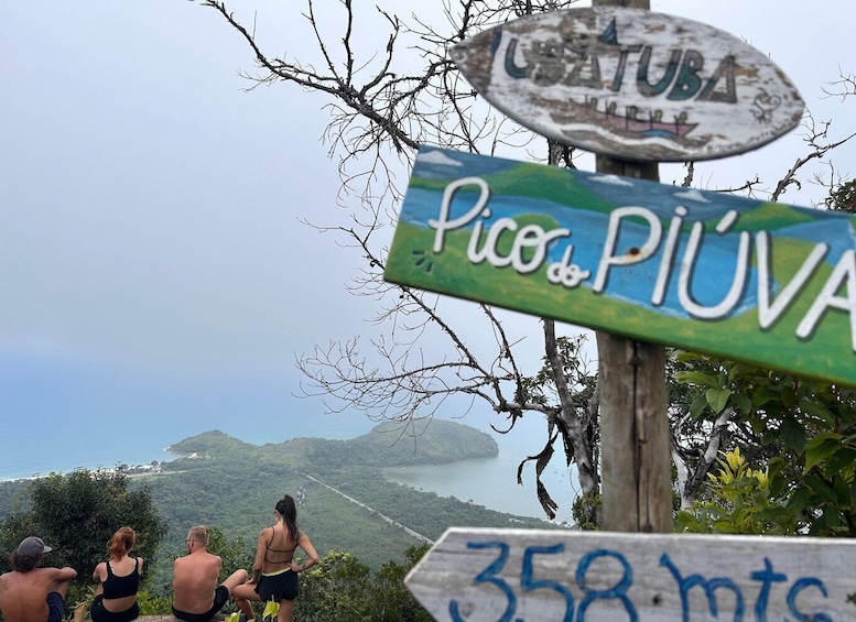 A signpost with 'Pico de Piúva' and 'Ustuba' written on it, overlooking a scenic landscape with a body of water and hills. 1