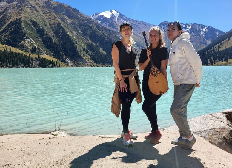 Three people standing by a lake with mountains in the background. 1