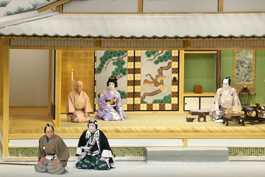 A traditional Japanese stage with four actors in period costumes, a wooden structure, and a green wall with a painting. 1