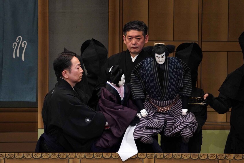 A person in traditional Japanese attire sitting on a stage with a backdrop of wooden paneling and a banner with Japanese characters. 4