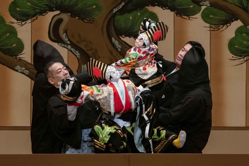 A person in traditional Japanese attire with a colorful costume, being carried by two individuals in black costumes. 5