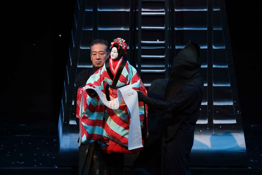 A person in traditional Japanese attire holding a scroll, standing in front of a backdrop with a grid pattern. 3
