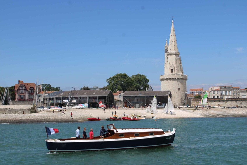 A boat with people on a calm water body near a sandy beach with buildings and a tower in the background. 1