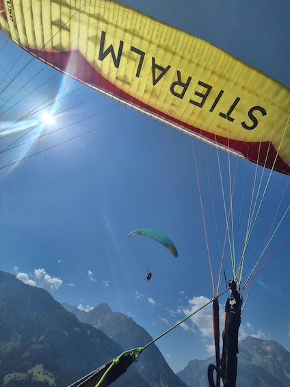 A paraglider with a yellow and red parachute labeled 'STEFAN' against a clear blue sky, with mountains in the background. 3