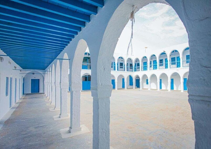Un couloir aux murs blancs et aux touches de bleu, qui mène à une cour intérieure dotée d’arcades et d’un toit bleu. 1