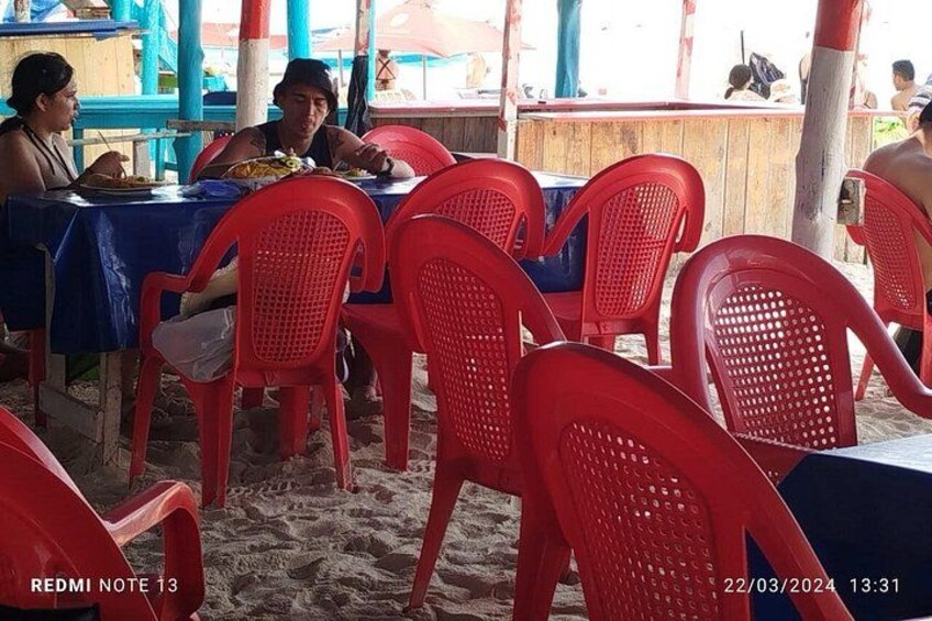A beachside restaurant with red plastic chairs and tables, covered by a blue awning. 2
