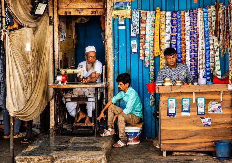 A street vendor stall with a sewing machine, a man sitting on a bucket, and various packaged goods hanging on the wall. 2